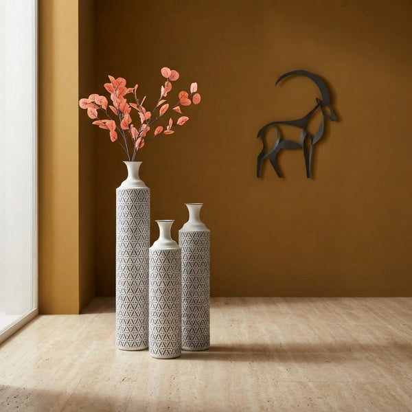 Three modern cylindrical metal vases in white and charcoal arranged on a stone floor in a minimalist room with warm lighting.
